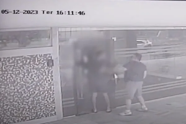 Imagem em preto e branco. Print de câmera de segurança mostra momento em que homem de camiseta escura, bermuda clara e tênis branco agride outro homem, que não tem o rosto nítido, mas veste camiseta e bermuda escuras. Os dois estão próximos a uma portaria de vidro