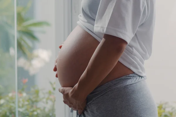 Foto colorida de mulher grávida com a mão na barriga em vrente a uma estrutura de vidro - Metrópoles