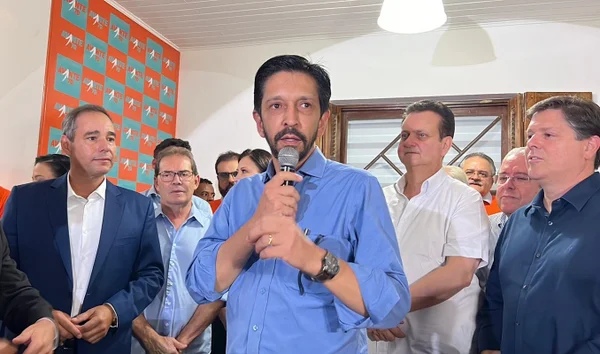 Imagem colorida mostra Ricardo Nunes, homem branco, de camisa azul, cabelo e barba pretos, segurando um microfone com as duas mãos, cercado de um grupo de homens brancos de meia idade - Metrópoles