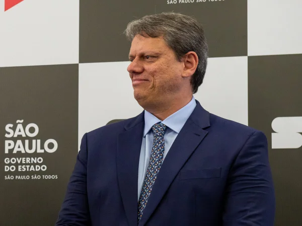 Imagem colorida mostra Tarcísio de Freitas, homem branco e grisalho, de terno e gravatas azuis, em imagem do peito para cima, olhando para seu lado direito e sorrindo, à frente de um banner quadriculado preto e branco - Metrópoles