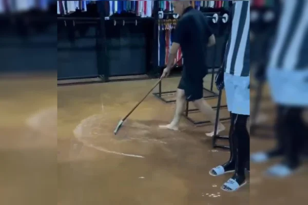Imagem colorida de um homem branco, com camiseta e bermuda preta, arrastando o chão encharcado de uma loja com um rodo. Ao lado, há um manequim