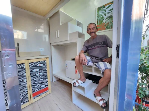 imagem colorida mostra homem sentado em móvel branco. ele veste uma camiseta cinza e um shorts claro. metrópoles