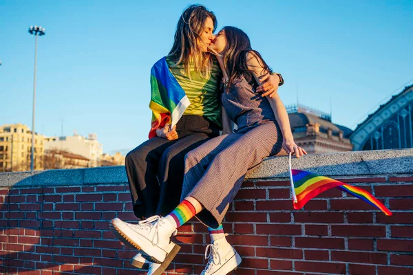 Foto colorida de um casal de mulheres sentadas em cima de um muro se beijando e sorrindo com bandeiras coloridas nas mãos - Metrópoles