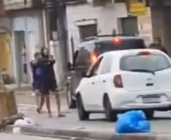 Imagem de homem com blusa de capuz apontando arma em direção a carro branco - Metrópoles