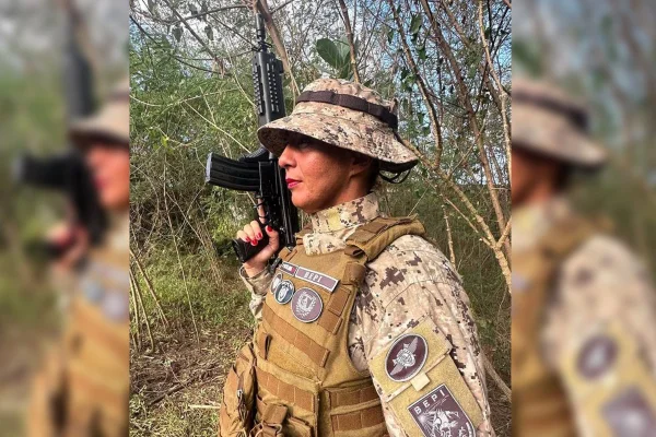 Foto de uma mulher de perfil, vestindo farda de cor areia e segurando arma de fogo para o alto na mão direito, em um ambiente com vegetação natural