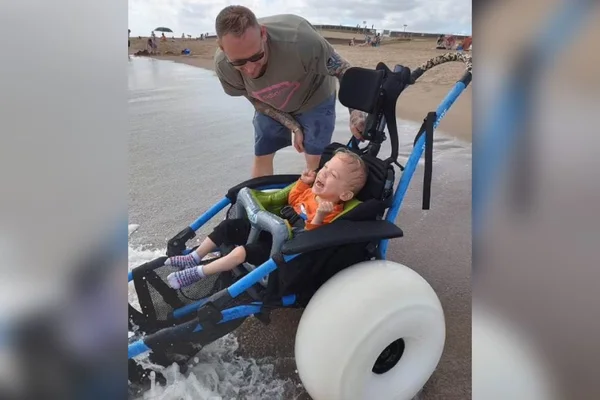 fotografia de criança com paralisia cerebral em uma cadeira de rodas especial se divertindo na praia ao lado do pai