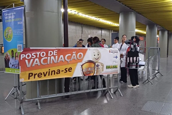 Imagem colorida com cartaz "Posto de Vacinação - Previna-se" dentro de estação de trem; pessoas formam fila atrás - Metróopoles