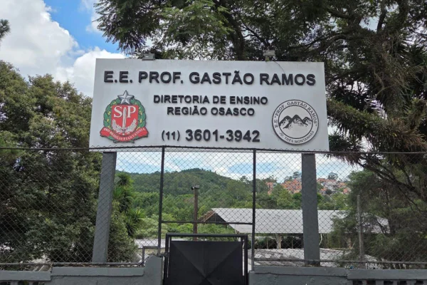 imagem colorida mostra placa com nome da escola estadual professor gastão ramos. ao redor da placa é possível observar uma construção e vegetação verde