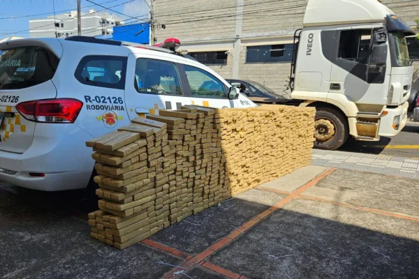 Imagem colorida mostra tijolos de maconha empilhadas, ao lado de viatura da PM. Caminhão aparece atrás - Metrópoles