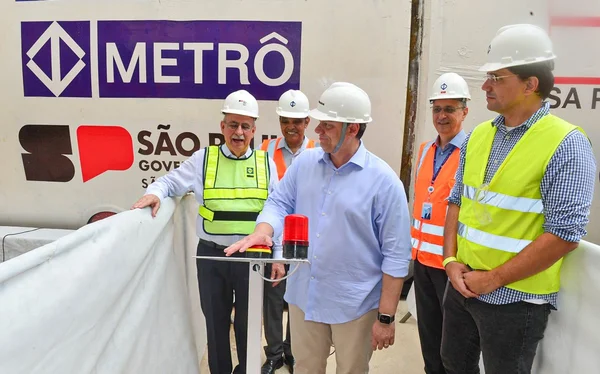 Imagem colorida mostra Tarcísio de Freitas e mais pessoas, vestidas com roupas sociais, capacete de obra e coletes refletores, durante visita a obras do metro. Todos são homens brancos de meia idade. Tarcísio está de frente para um púlpito transparente - Metrópoles