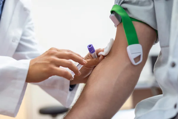 Preparação de exame de sangue por médica, uniforme médico, mesa, sala branca e iluminada; enfermeira perfura a veia do braço do paciente com agulha, tubo em branco. Metrópoles