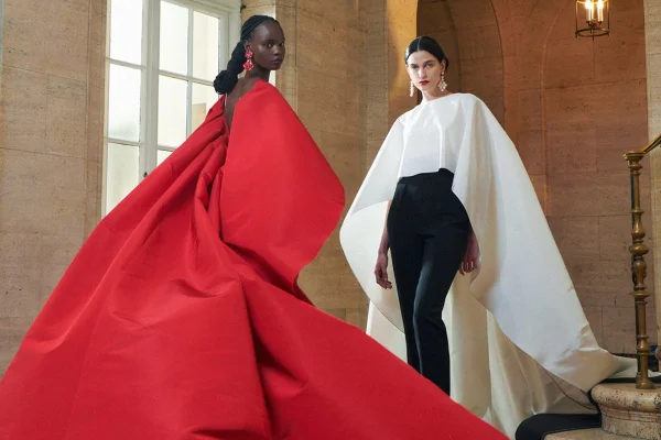 Duas modelos jovens posam para foto de campanha da marca Carolina Herrera. A primeira é negra, possui o cabelo castanho trançado e usa um vestido longo e grande vermelho. A segunda é branca, possui o cabelo liso e usa uma blusa branca com babado e uma calça preta. - Metrópoles.