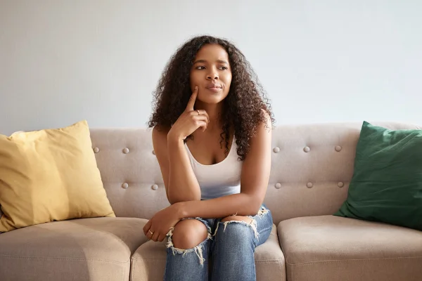Imagem de garota com aparência africana sentada isolada em um sofá cinza com almofadas amarelas e verdes, com um olhar pensativo e duvidoso, tocando o queixo. Metrópoles