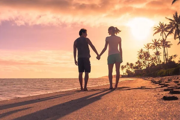 Casal andando de mãos dadas na praia ao por do sol - Metrópoles