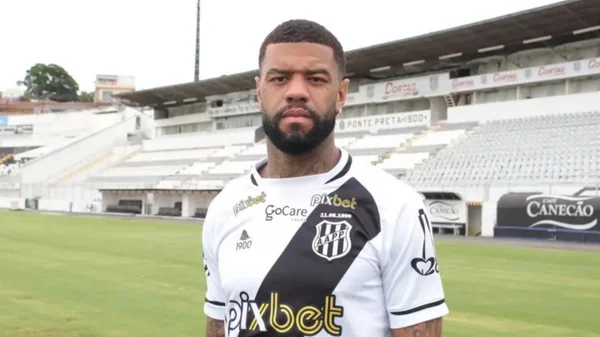 Imagem colorida mostra o lateral-esquerdo Junior Tavares, um homem negro, de barba, vestido com a camisa da Ponte Preta, que é branca com uma faixa transversal preta - Metrópoles
