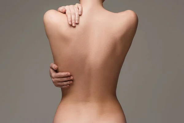 Foto colorida em close de uma mulher nua e de costas, com as mãos no ombro e na costela, se abraçando, em um fundo cinza - Metrópoles