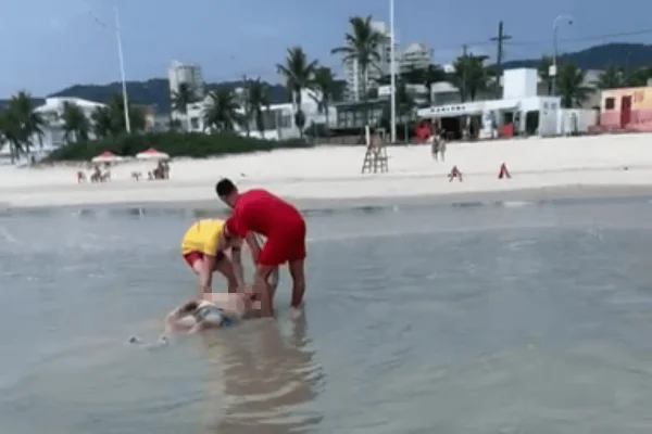 Imagem colorida de um homem branco morto sendo retirado do mar por um bombeiro e outro banhista - Metrópoles