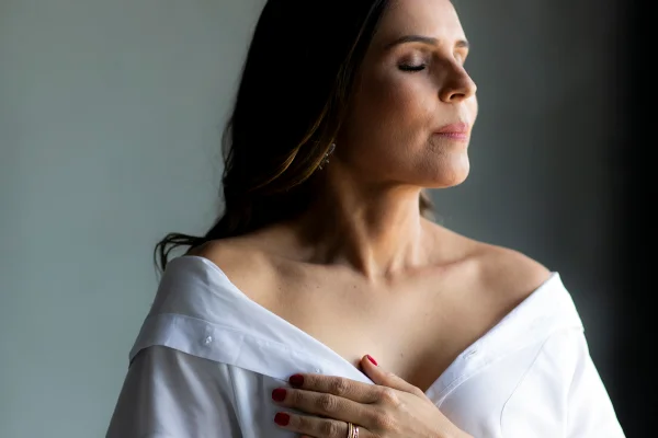 Foto colorida de mulher com blusa branca e mão em cima de um dos seios - Metrópoles.