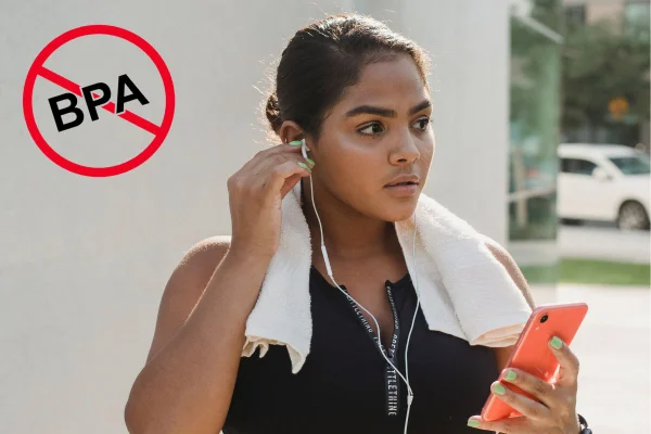 Mulher jovem com fones de ouvido, toalha branca no pescoço e celular na mão olhando para a lateral em uma rua - Metrópoles