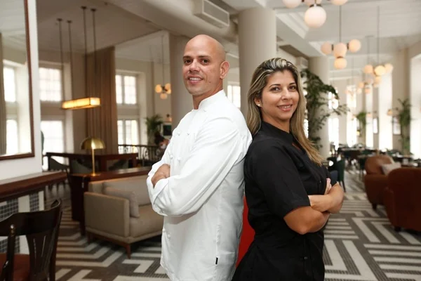 Um chefe de cozinha vestido de branco está de costas para a outra chefe de cozinha vestida de preto. Ambos estão sorrindo