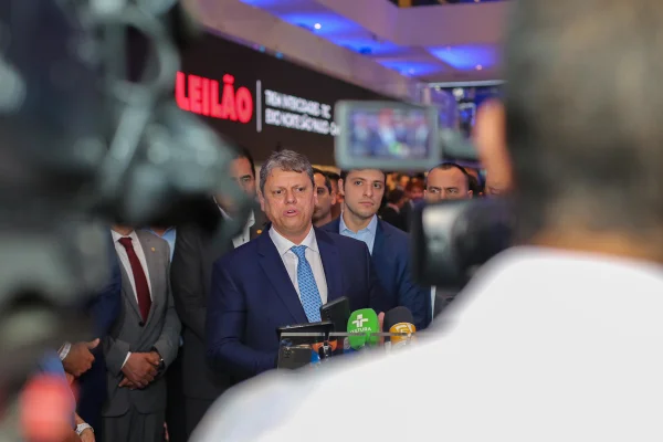 Imagem colorida mostra Tarcísio de Freitas, homem branco, grisalho, vestindo terno azul, camisa branca e gravata azul, entre câmeras de TV e rodeado de apoiadores, dando uma entrevista sobre leilão - Metrópoles