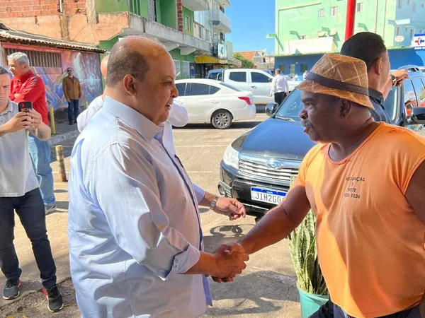 Homem de blusa social azul, calvo, apertando a mão de um homem negro, de blusa laranja e chapéu de palha