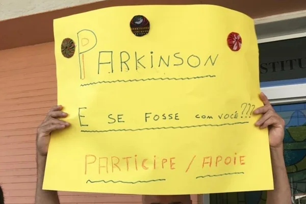 cartaz da cor amarela é erguido por uma pessoa. no papel, estão escritas perguntas sobre a doença de Parkinson