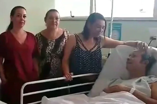 Imagem colorida de mulheres em pé ao lado de homem deitado em cama de hospital. Metrópoles