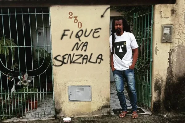 foto colorida de homem negro na frente do portão de casa ao lado de pichação racista