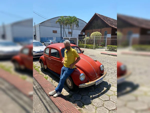 Homem de cabelos grisalhos, máscara e blusa amarela se encosta em carro vermelho estacionado em rua. Ao fundo, uma casa de madeira rústica e um ginásio