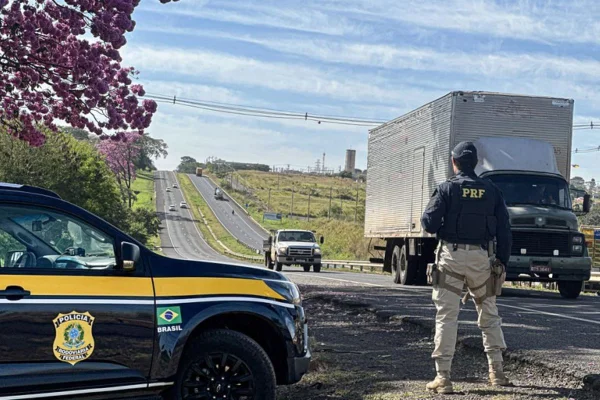 Imagens coloridas mostram agente da PRF parado nas margens de estrada durante fiscalização, viatura estacionada ao lado dele