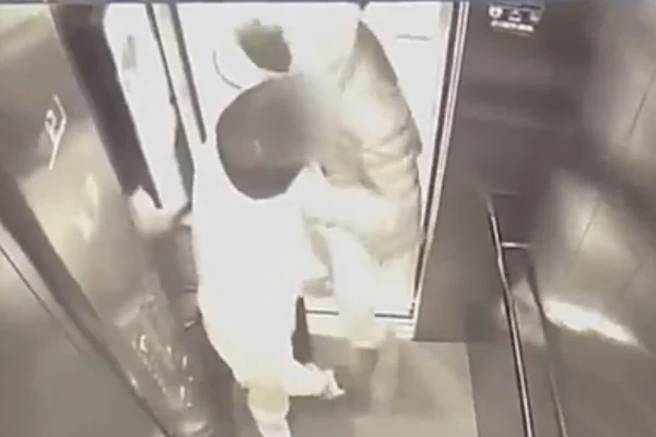 Imagem preto e branco de homem careca, de jaqueta preta, dando soco no rosto do rapaz mexendo no celular. Ambos estão em um elevador - Metrópoles
