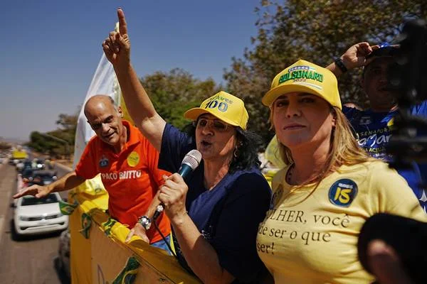 Fotografia colorida de duas mulheres de boné e um homem ao fundo de camiseta laranja