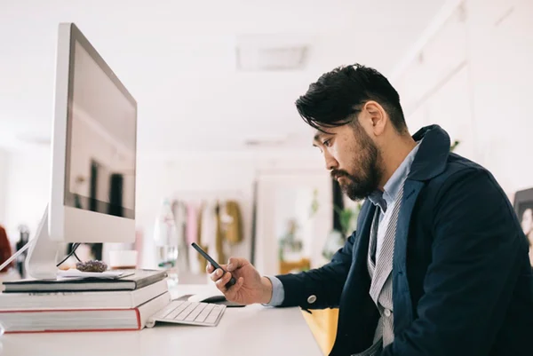 Imagem colorida: homem sentado em frente a computador mexendo no celular - Metrópoles