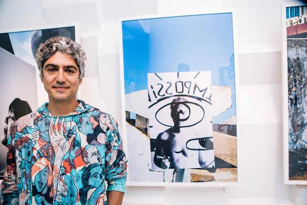 Homem jovem e branco, de cabelo ondulado grisalho, posando para foto no Museu da República em frente a um quadro que ele mesmo pintou