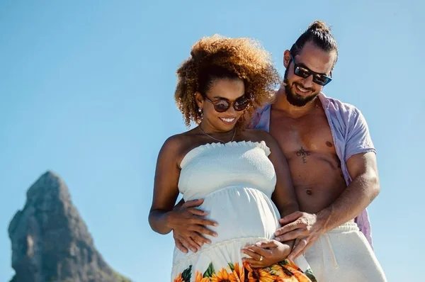 Foto colorida de Jeniffer Nascimento e do marido, Jean Amorim. Ela está de branco e mostra a barriga de grávida, enquanto ele está de camisa aberta - Metrópoles