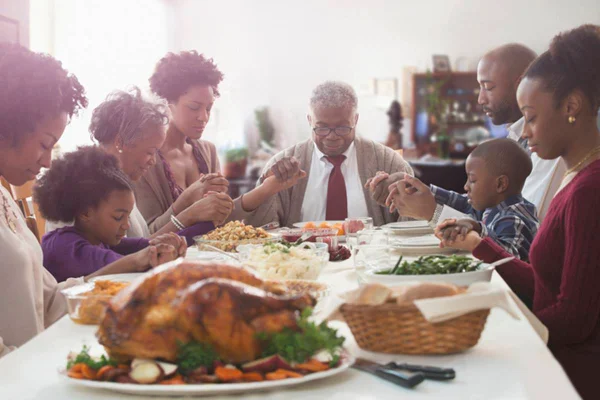 Na foto vemos uma família de pessoas pretas de mãos dadas em volta de uma mesa com ceia de natal