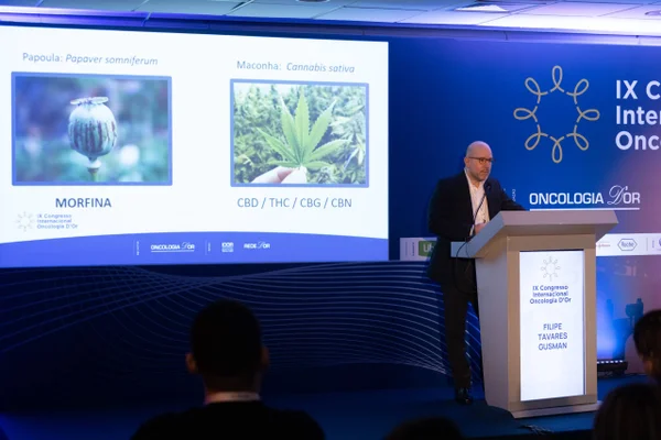 Foto mostra o médico Felipe Gusman em uma palestra. Uma TV ao seu fundo mostra imagens de plantas de morfina e de cannabis