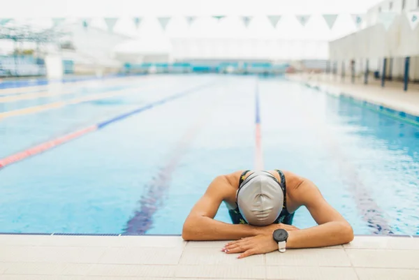 Mulher de touca encosta a cabeça sobre os braços na borda de uma piscina - Metrópoles