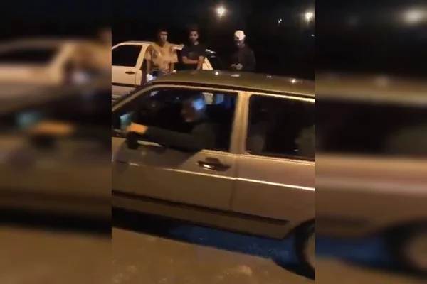 Homem dentro de carro com a mão na janela e outros três homens atrás, encostados em carro, à noite