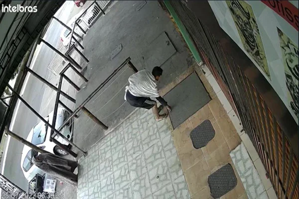 Homem está agachado em fosso enquanto mulher com coberta caminham em frente a estabelecimento