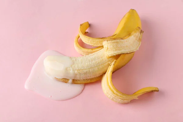 Foto colorida de uma banana meio descascada com um liquido branco na ponta em um fundo cor-de-rosa - Metrópoles
