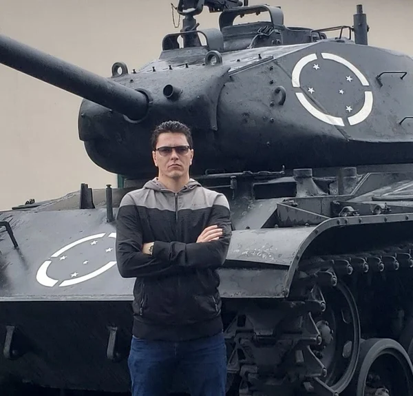 Carlos Victor Carvalho, conhecido como CVC, terceiro alvo preso em operação que investiga terrorismo bolsonarista no DF. Na imagem ele aparece sério, de braços cruzados com um tanque atrás - Metrópoles
