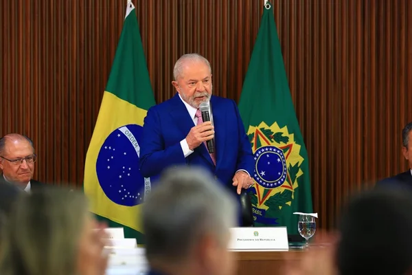 O presidente Lula faz primeira reunião ministerial no Palácio do Planalto e pede boa relação com o Congresso Nacional. Na imagem ele está de pé, na ponta de uma mesa, falando em microfone com bandeiras do Brasil atrás e ao lado de Alckmin - Metrópoles