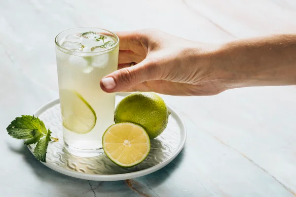 Imagem mostra em close-up uma mão colocando um copo com suco de limão em cima de uma pequena bandeija. Metrópoles