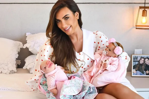 Imagem colorida de Nicole Bahls sentada na cama, carregando bonecas reborn