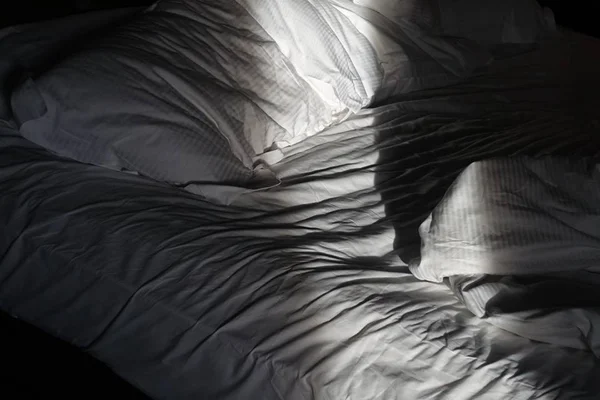 Foto colorida em close de uma cama com lençóis brancos bagunçados com um feixe de luz no travesseiro - Metrópoles