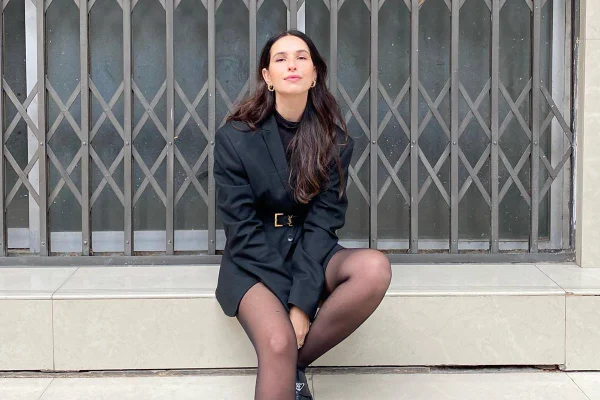 Vic Hollo usa blazer preto com meia calça preta encostada na parede - Metrópoles