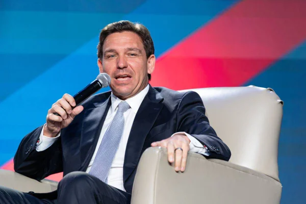 imagem colorida mostra ron desantis sentado em cadeira branca e segurando microfone, ele é branco com cabelos pretos e usa terno azul escuro