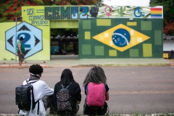 Três alunos sentados na calçada observando a porta da escola. Cor verde do muro da escola predomina na foto